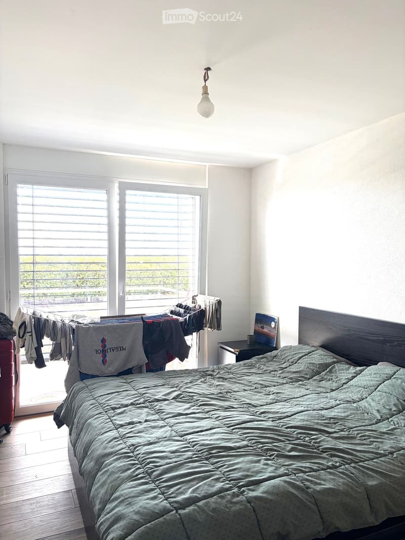 Bright bedroom with wooden floor, large windows with blinds, bed with green duvet, nightstand with picture frame, clothesline with hanging clothes, and a red suitcase near the window.