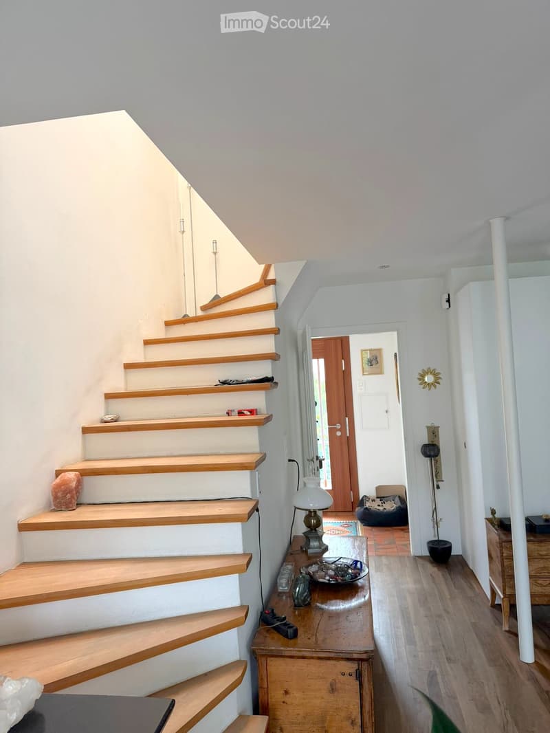 Staircase with wooden steps and white walls, leading to a hallway with a small table, lamp, and a pet bed visible in the background.