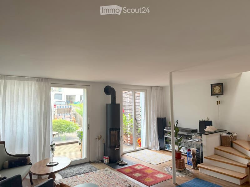 Bright living room with large windows and sliding doors leading to a patio, featuring a modern wood-burning stove, a grey sofa, a wooden coffee table, and several area rugs.