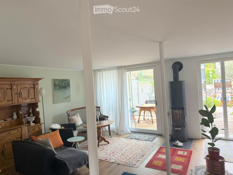 Living room with a grey sectional sofa, coffee table, armchair, side table, floor lamp, wooden cabinet, painting, fireplace, and outdoor table and chairs visible through large glass doors.