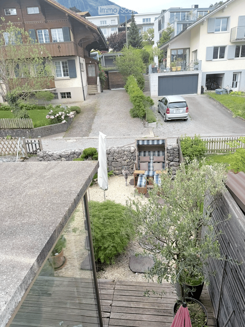 A small, well-maintained garden area with a wooden deck, potted plants, and a stone pathway leading to a seating area with a lounge chair and small table.