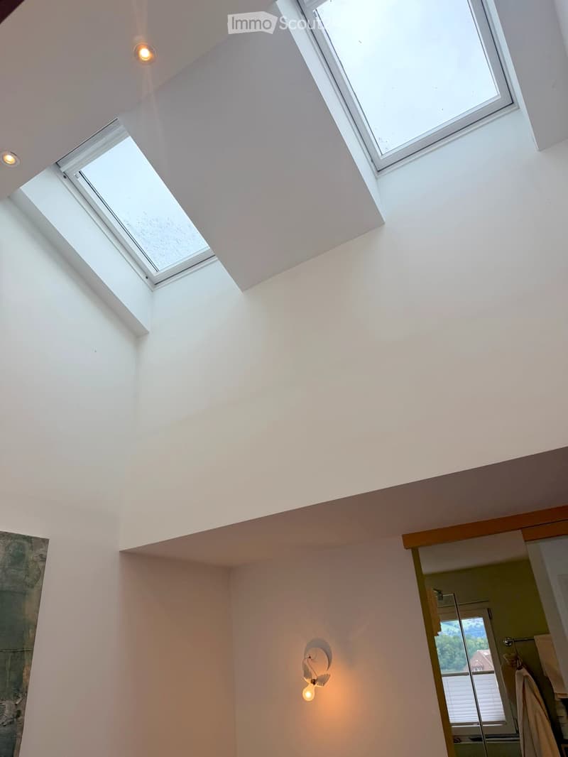 Bright, modern room with white walls and a high, slanted ceiling. Features three skylights and a wall-mounted light fixture. A doorway leads to a bathroom with a visible towel and window.