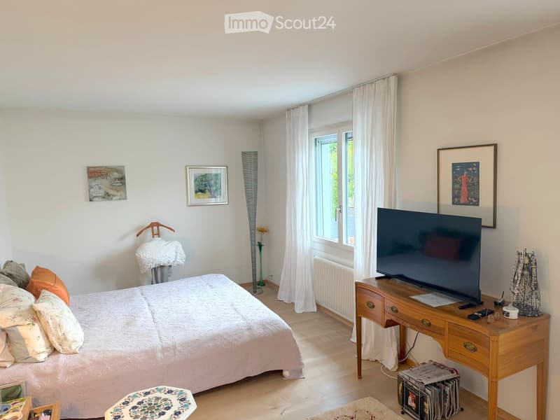 Bright bedroom with a double bed, nightstand, wooden table, and television. Features large windows with white curtains, artwork on the walls, and a small seating area with a stool.