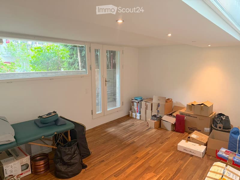 Room with wooden flooring, large window, sliding glass door, massage table, storage boxes, and bags.