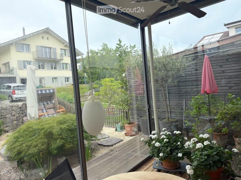 A well-maintained garden area with wooden decking, potted plants, a small tree, and a pink umbrella. The space is enclosed by glass walls, with a view of neighboring houses and greenery.