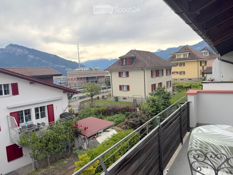 Balcony overlooking a residential area with houses, greenery, and distant mountains. Features a table and chairs, metal railing, and a view of the surrounding landscape.