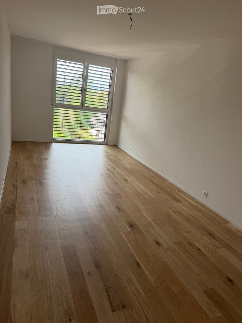 Empty room with light wooden flooring and white walls, featuring a large window with blinds and a door leading outside.