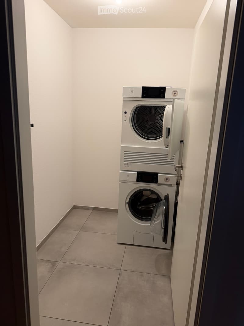 Modern bathroom with a stacked washing machine and dryer unit, white tiled walls, and a tiled floor.
