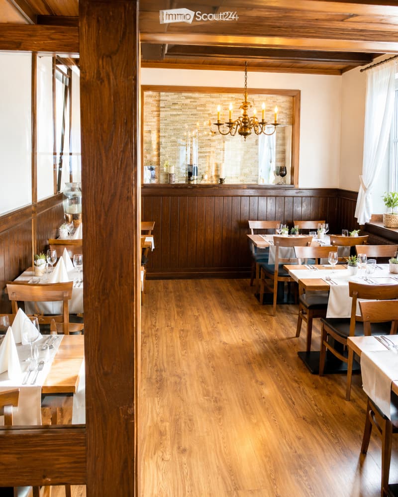 Elegant restaurant interior with wooden floors, tables set with white linens, wine glasses, and silverware, wooden chairs, a chandelier, and a brick accent wall with a mirror and decorative items.