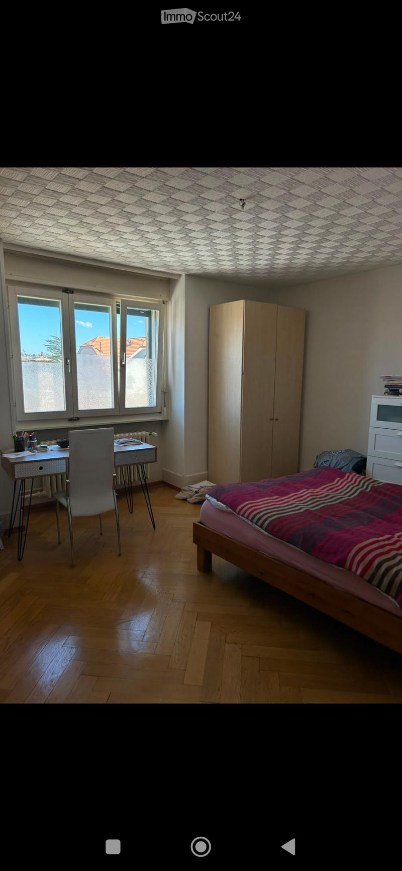 Bright bedroom with wooden flooring, large windows, a bed with striped bedding, a desk with a chair, and a wardrobe.
