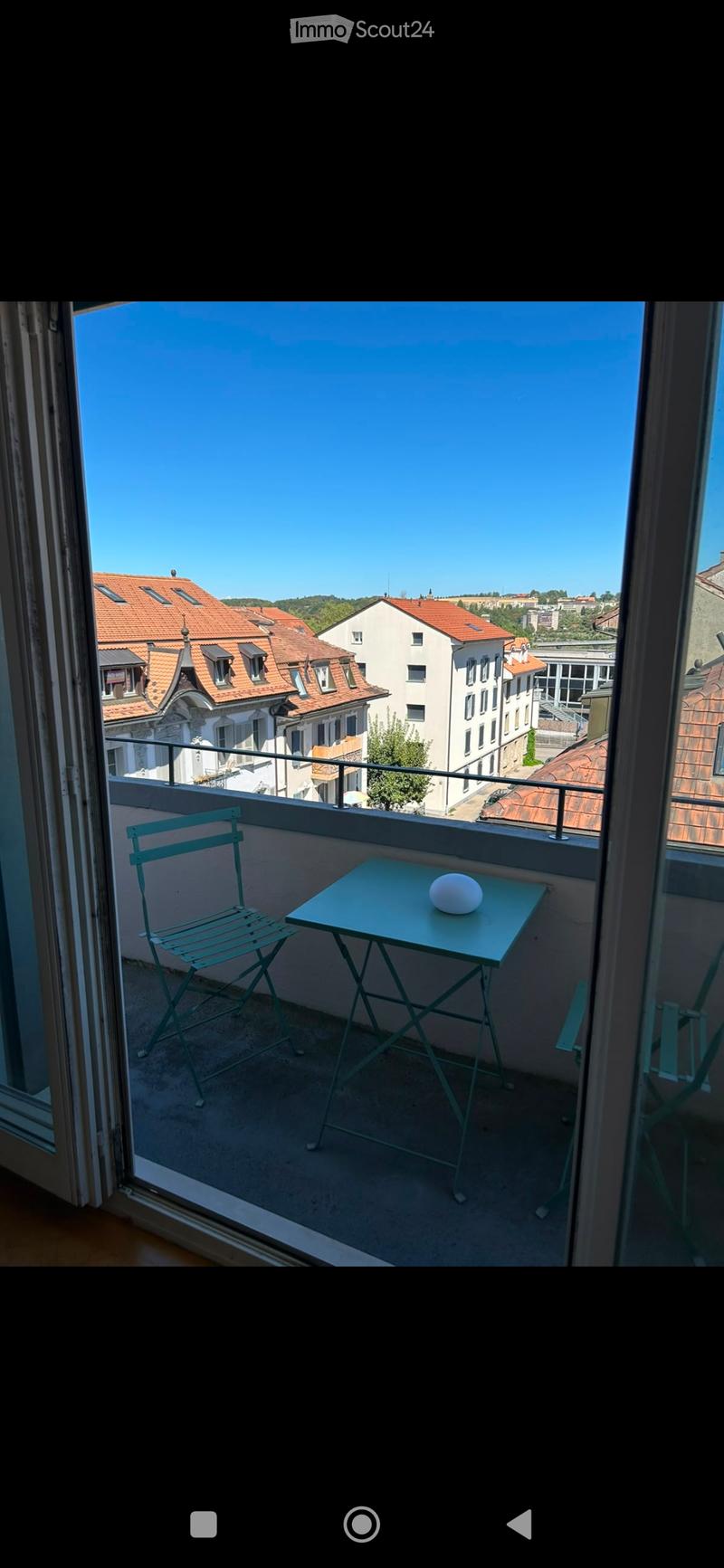 Balcony with a small table and a chair, overlooking a cityscape with red-tiled roofs and buildings.