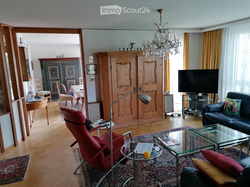 Spacious living room with wooden flooring, a red leather chair, a glass coffee table, a black leather sofa, a chandelier, and a dining area with a table and chairs.