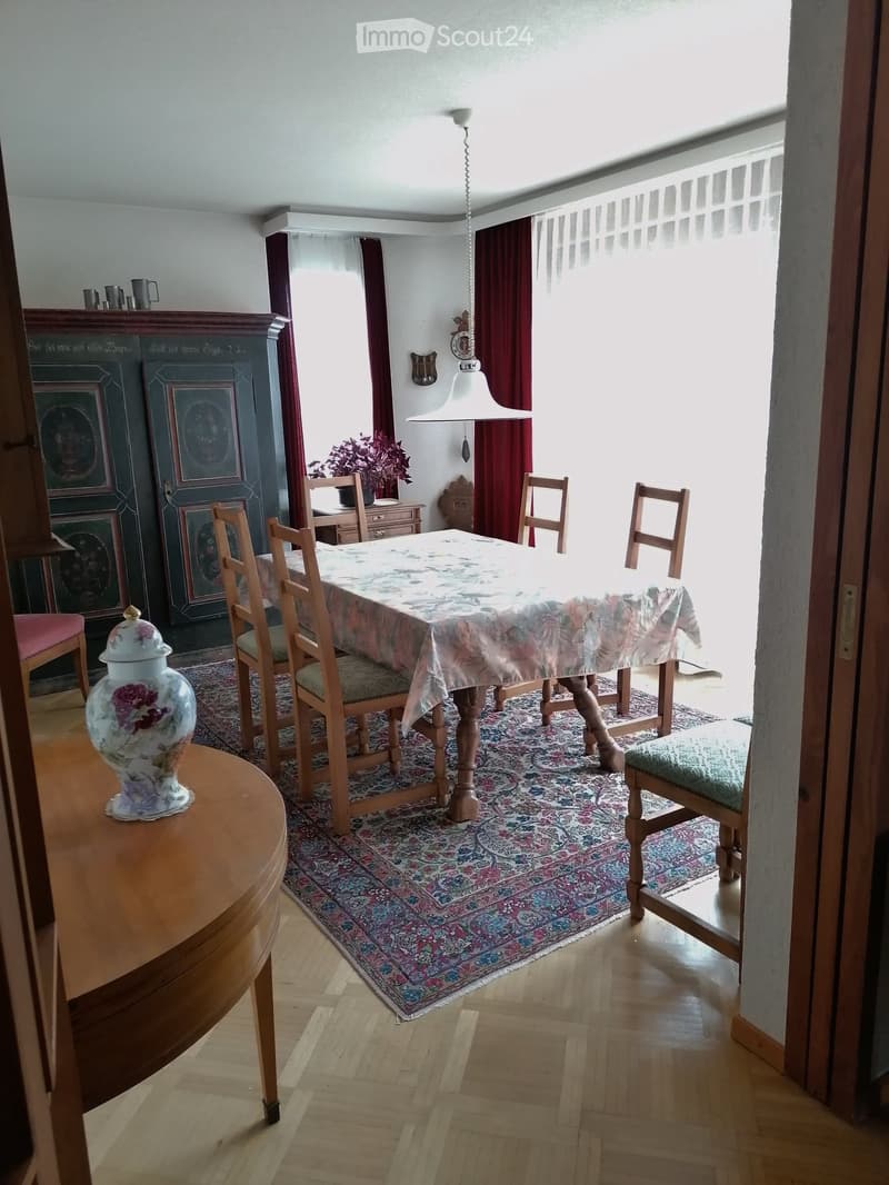 Dining room with wooden floor, patterned rug, dining table with floral tablecloth, six wooden chairs, decorative vase, cabinet, and hanging lamp.