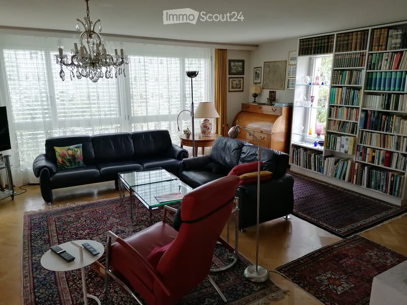 Spacious living room with wooden flooring, large windows, a black leather sofa, red armchair, glass coffee table, bookshelf, and decorative lamps.