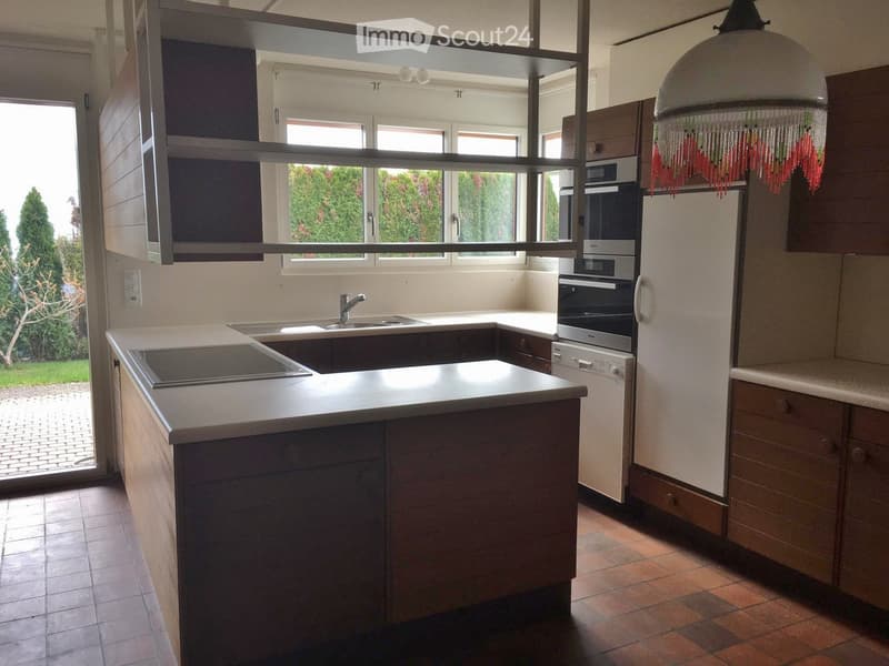 Modern kitchen with a central island, sink, oven, refrigerator, dishwasher, and cabinets. The space features a tiled floor and large windows offering a view of greenery outside.