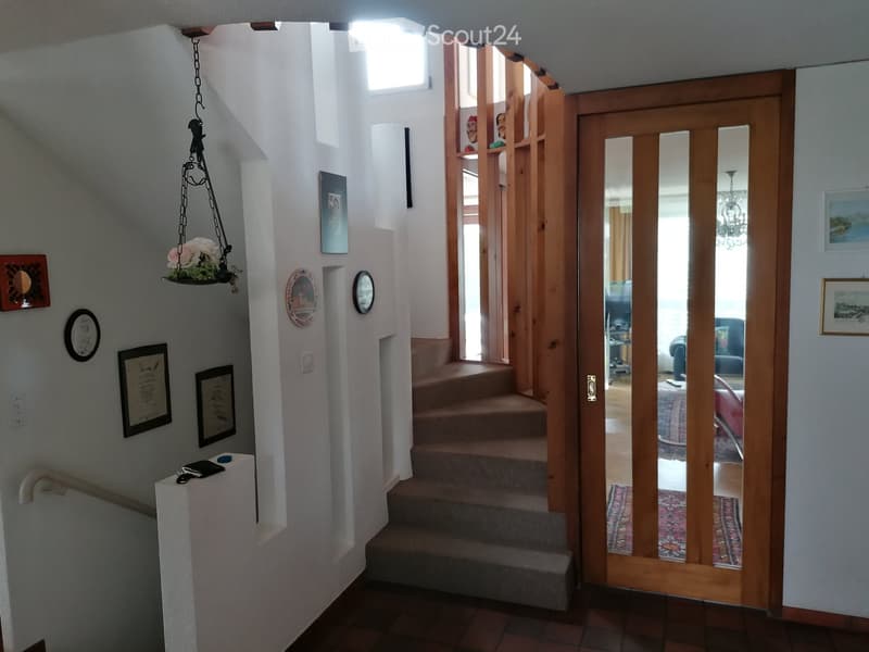 Staircase leading to an upper level with wooden railings, framed artwork on walls, and a hanging flower basket. A glass door opens to a room with visible furniture.