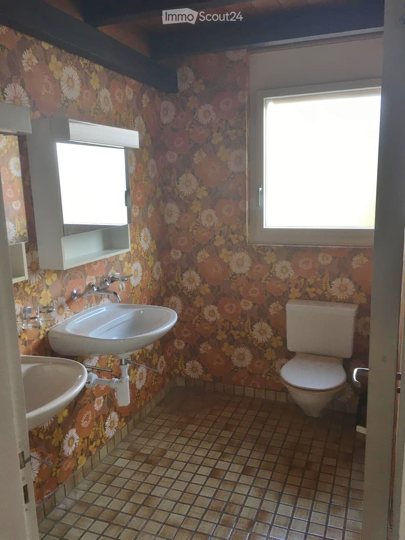 Bathroom with floral wallpaper, two sinks, a toilet, and a mirror with a shelf above it. The room has tiled flooring and two windows.