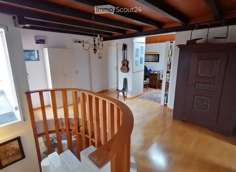 Spacious hallway with wooden flooring, curved staircase with wooden railing, white walls, and several doors leading to different rooms. Visible items include a guitar on the wall, a small chair, a cabinet, and a chandelier hanging from the ceiling.