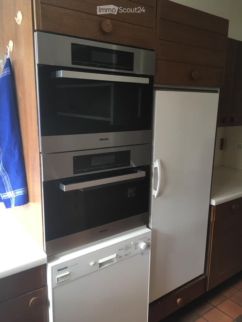 Modern kitchen with stainless steel appliances including a dishwasher, refrigerator, and built-in oven, wooden cabinets, and tiled floor.