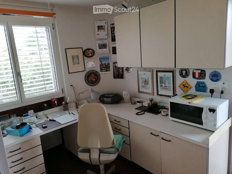 A well-organized home office with a white desk, a beige office chair, a microwave, a lamp, and various office supplies. The walls are decorated with framed pictures, calendars, and a few decorative plates.