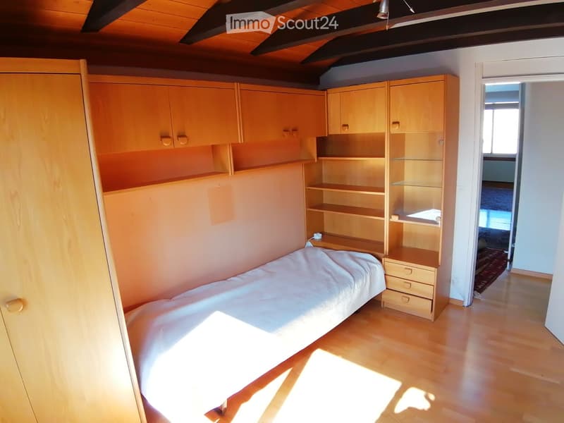 Cozy bedroom with a single bed, built-in wardrobe, shelves, and a nightstand. The room features wooden flooring and warm lighting.