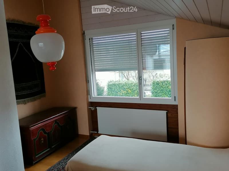 Bedroom with a bed, a wooden cabinet, a radiator, and a hanging lamp. The room has wooden flooring and a window with blinds.