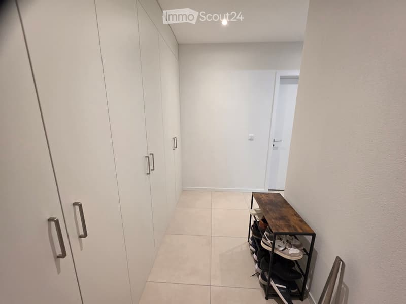 A narrow hallway with white walls, tiled floor, built-in wardrobes, and a small wooden table with a shoe rack holding multiple pairs of shoes.