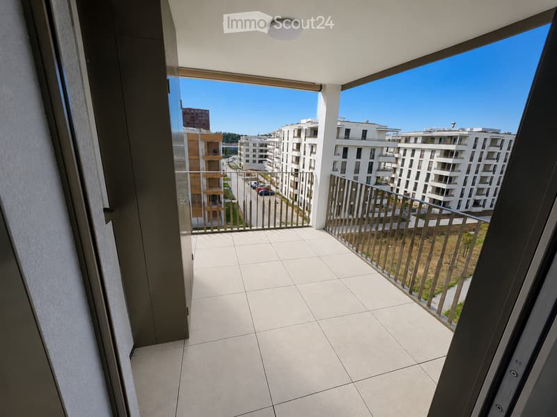 Modern balcony with tiled floor, metal railing, and expansive view of a residential area with contemporary buildings and clear blue sky.