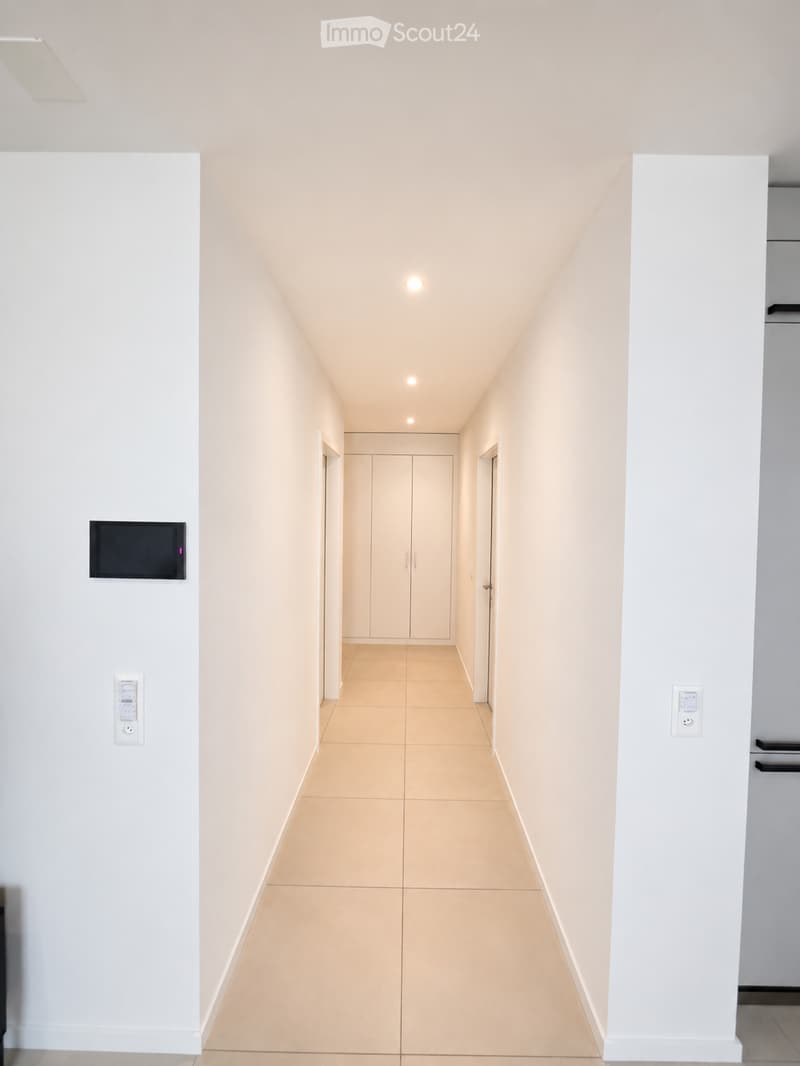 A clean, modern hallway with light-colored walls and tiled flooring, featuring recessed ceiling lights and a closed door at the end.