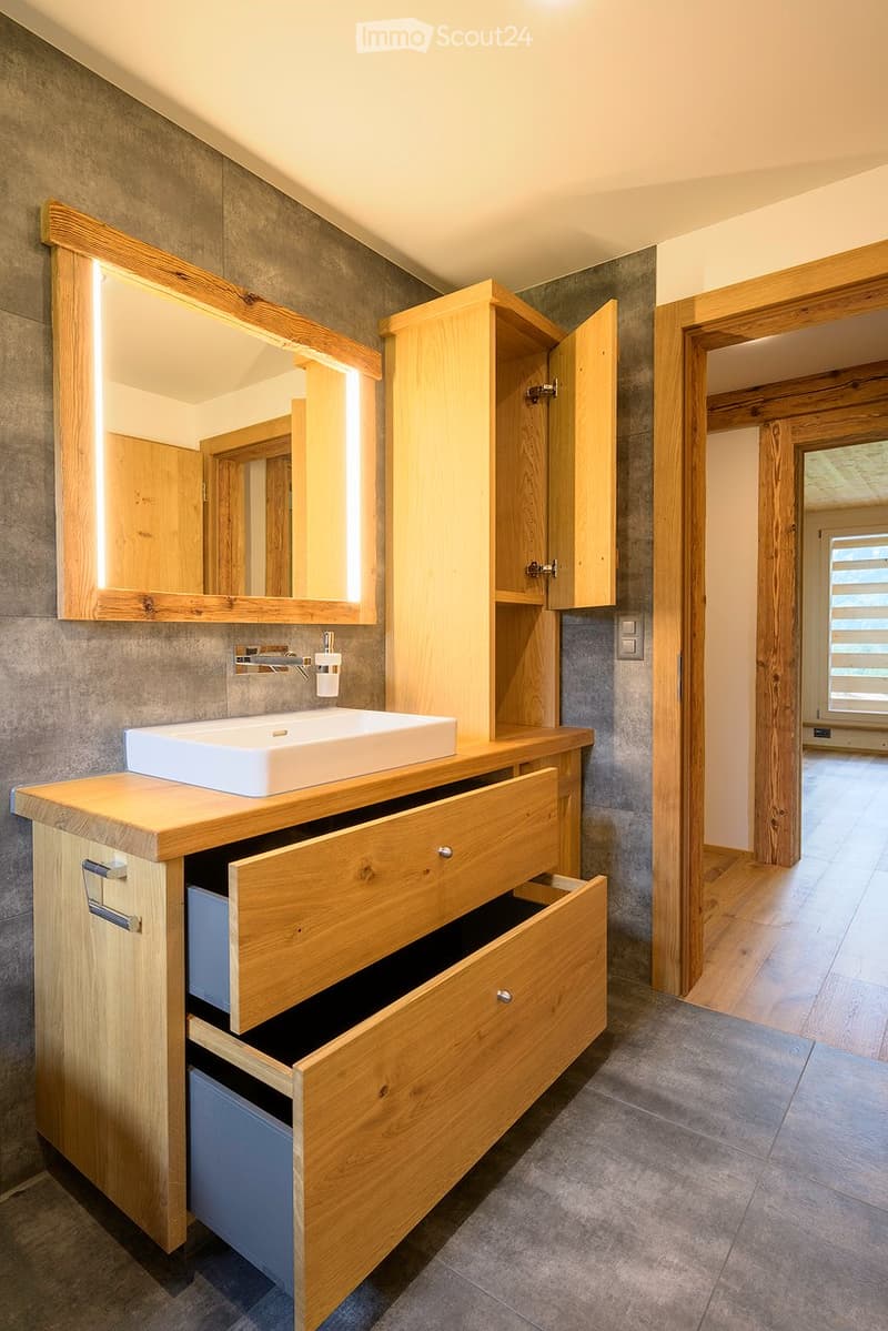 Modern bathroom with a wooden vanity, white sink, illuminated mirror, and open wooden cabinetry.