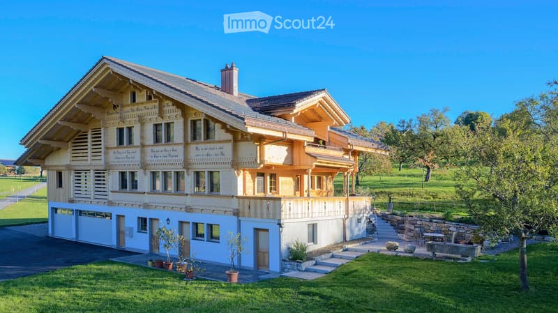 A large, traditional Swiss-style house with a prominent wooden facade, multiple windows, and a spacious balcony. The house features a well-maintained lawn, potted plants, and a stone staircase leading to the entrance.