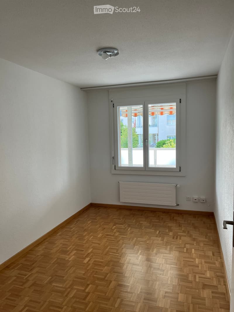 Empty room with wooden herringbone flooring, white walls, a large window with blinds, a radiator, and a ceiling light fixture.