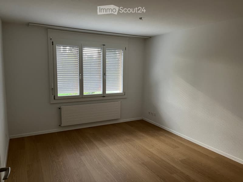 Empty room with wooden flooring, white walls, and a large window with blinds. A radiator is visible below the window.