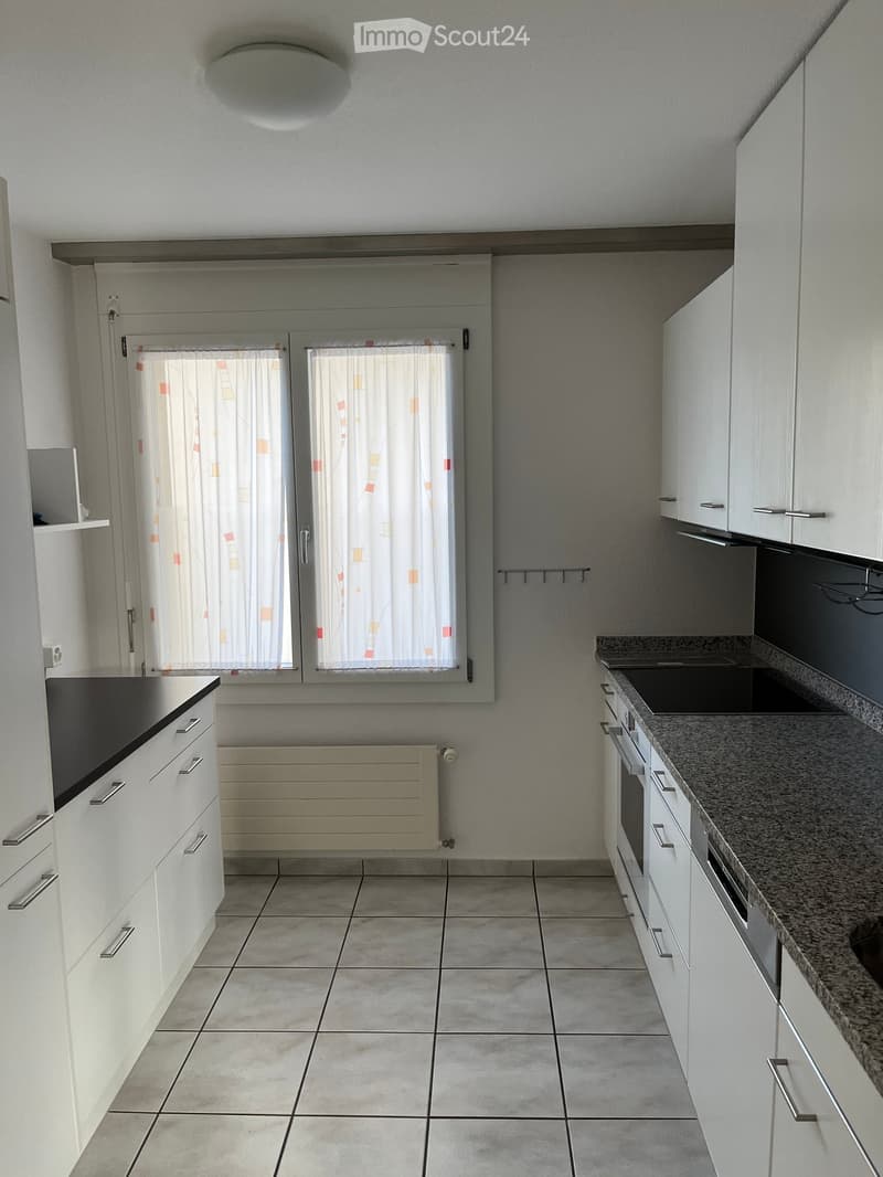 Modern kitchen with white cabinets, granite countertop, built-in oven, and tiled floor. Features a window with patterned curtains and a radiator below.