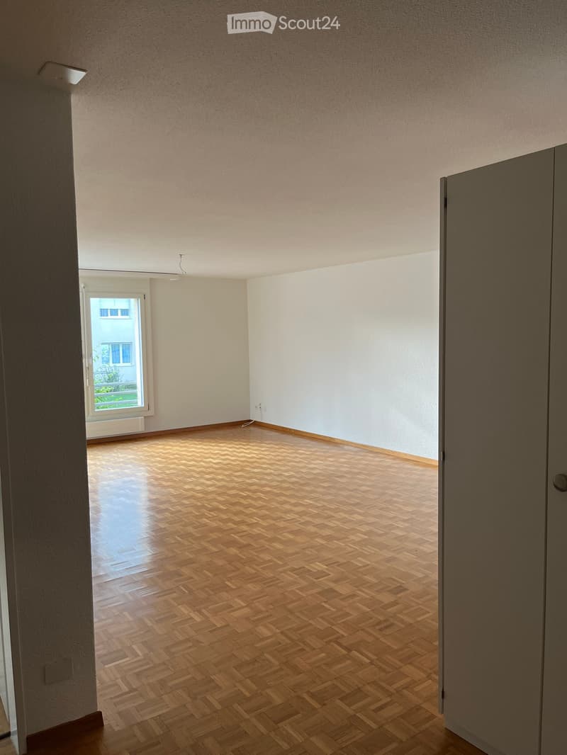 Empty room with wooden herringbone flooring, white walls, and a window with a view of a building outside.