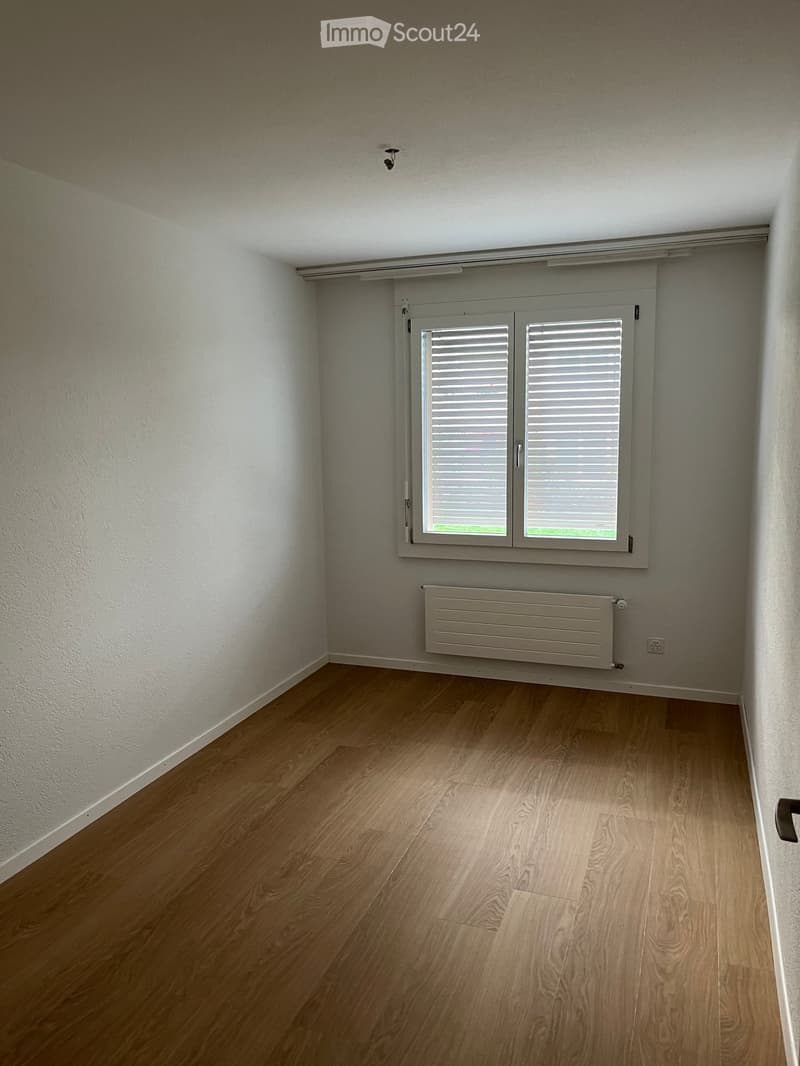 Empty room with light wooden flooring, white walls, a window with shutters, and a radiator below the window.