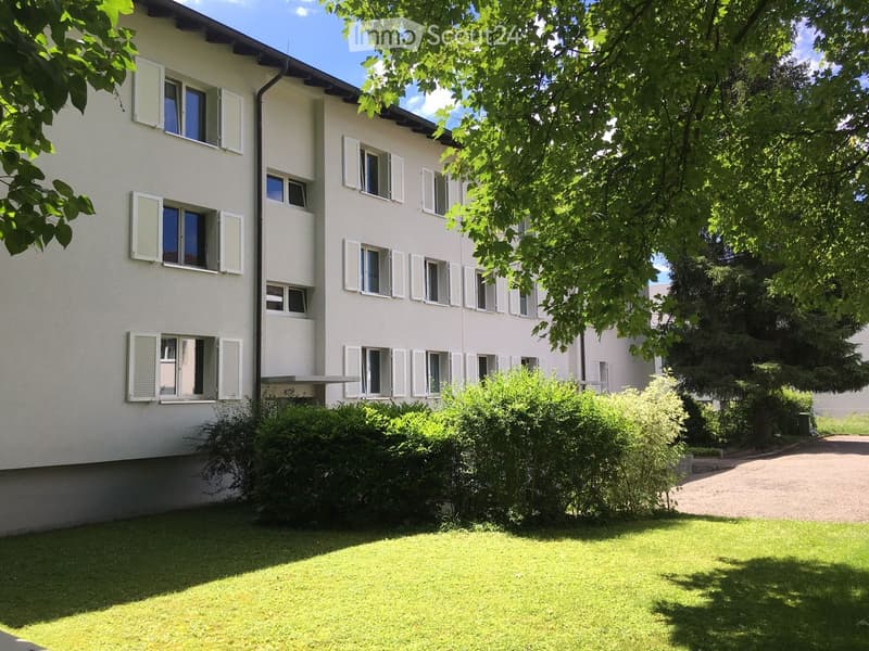 A multi-story residential building with white walls, multiple windows with shutters, surrounded by greenery and a well-maintained lawn.