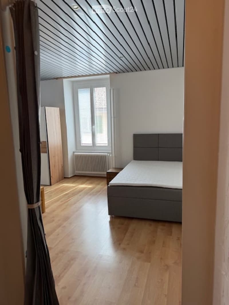 A small bedroom with wooden floor, a bed, a window with blinds, a wooden cabinet and a radiator below the window.
