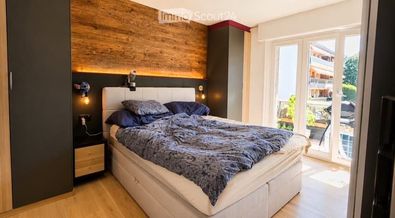 Modern bedroom with a large bed, nightstand, wall-mounted lamp, and decorative wall piece. The room features wooden flooring and a large window with a view of the outdoor area.