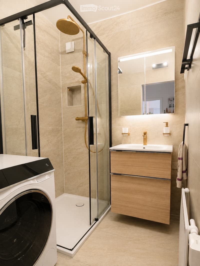 Modern bathroom with a glass-enclosed shower, gold fixtures, a wooden vanity with a sink, a large mirror, and a washing machine.