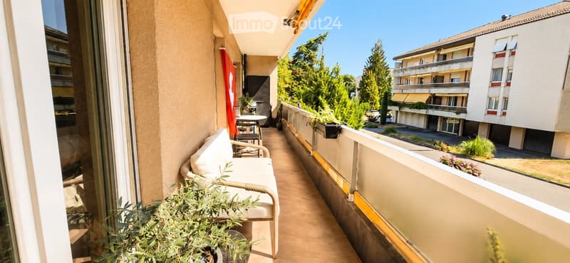Balcony with a white bench, small table, and potted plants, overlooking a residential area with trees and neighboring buildings.