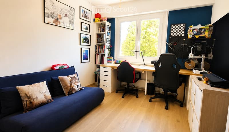 A modern, multi-functional room with a blue sofa, a desk with a chair, a bookshelf, and a large window with a view of greenery.