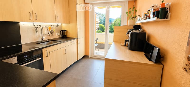 Modern kitchen with light wooden cabinets, stainless steel appliances, and a breakfast bar. Features a sink, oven, coffee maker, microwave, and a small chair near the sliding glass door.