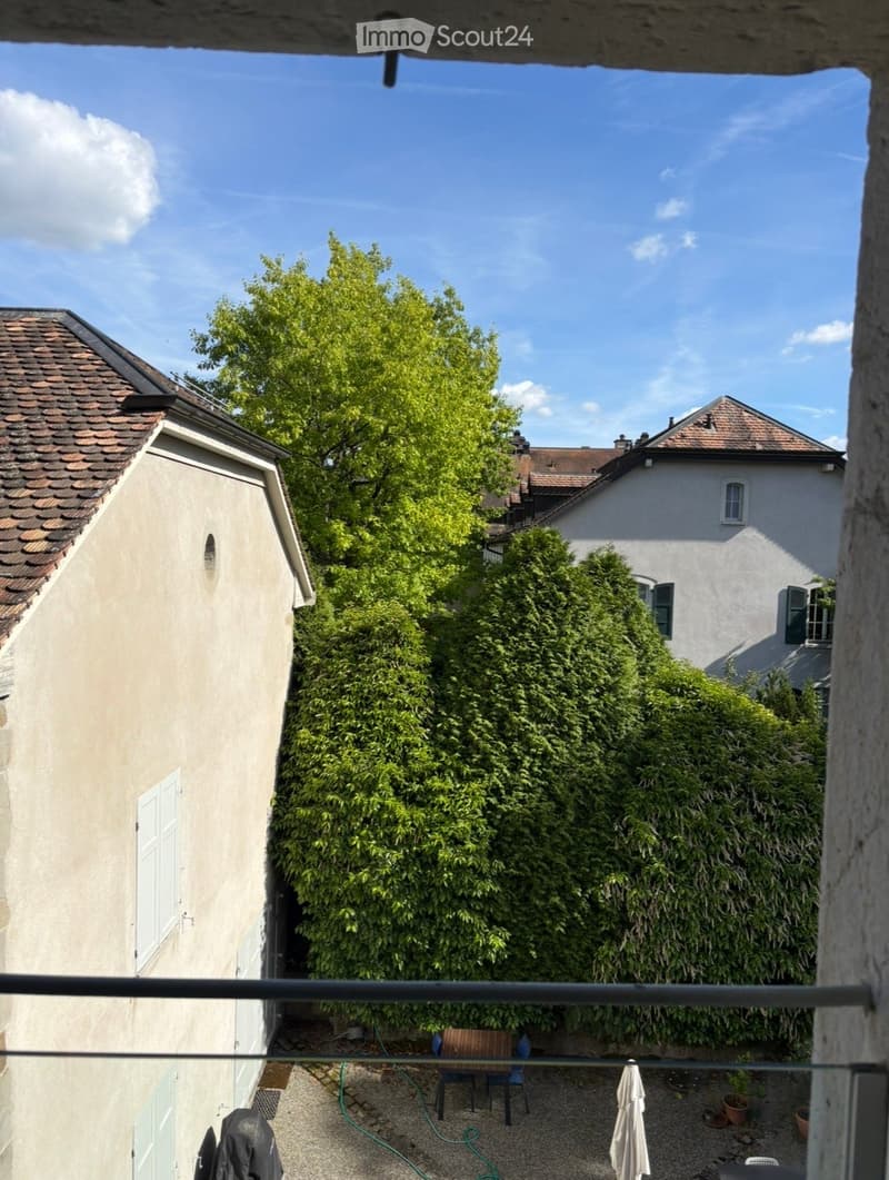Balcony overlooking a garden area with a table, chairs, and an umbrella, surrounded by lush greenery and neighboring buildings.