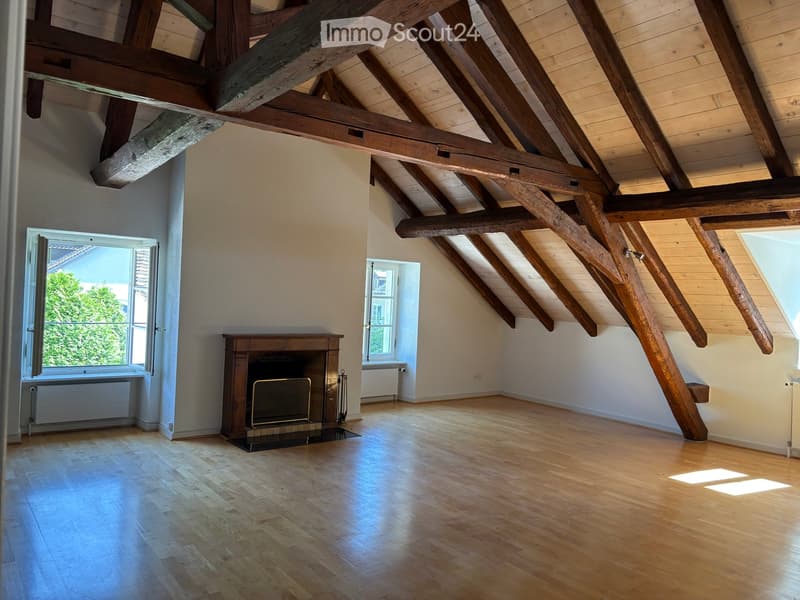 Empty attic room with exposed wooden beams, wooden flooring, and a fireplace with a glass front. Two windows provide natural light.