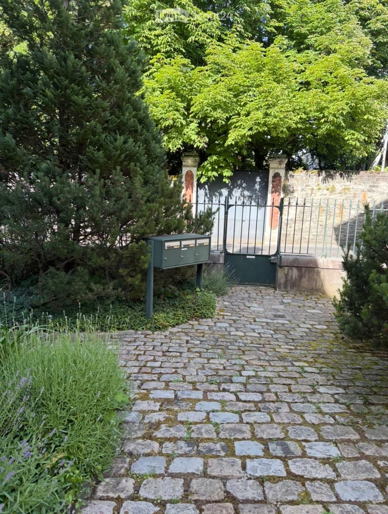 A cobblestone pathway leads to a gated entrance surrounded by lush greenery and trees. A green mailbox is positioned on the left side of the pathway.
