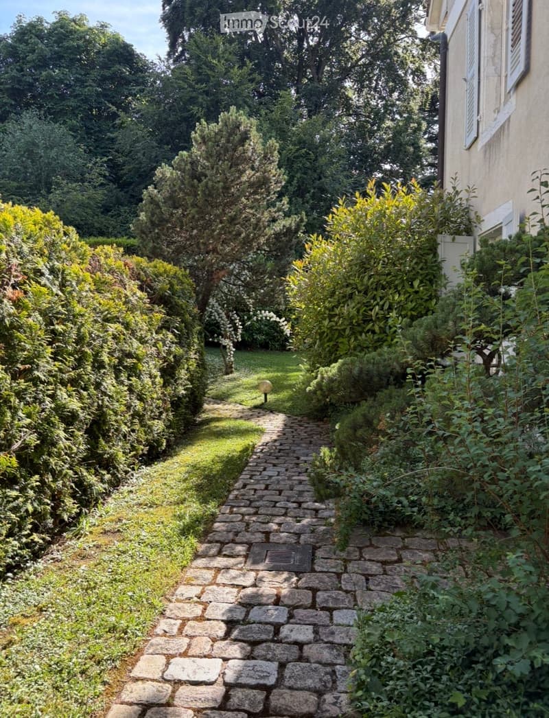 A well-maintained garden with a stone pathway leading through lush greenery and shrubs, bordered by a house on one side.