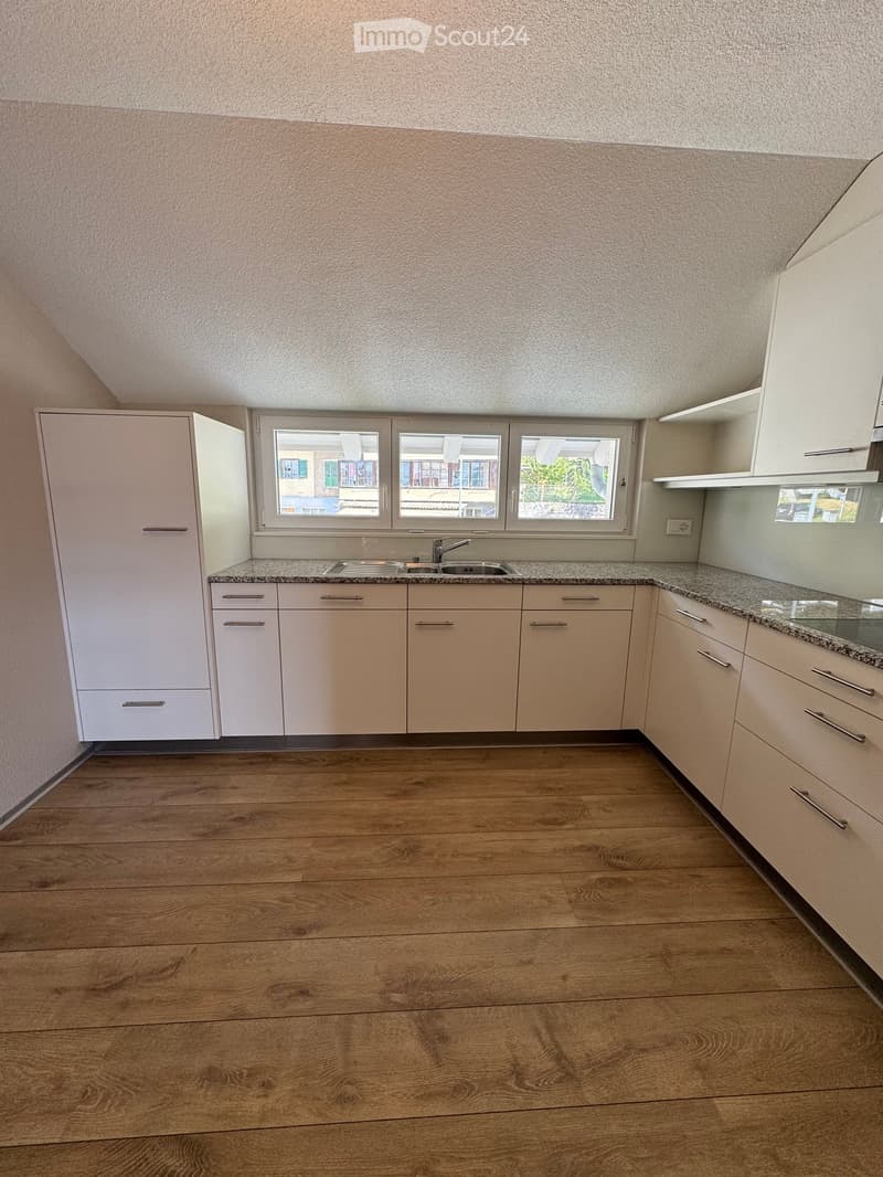 Empty kitchen with light-colored cabinets, granite countertop, and wooden flooring. Three windows provide natural light.