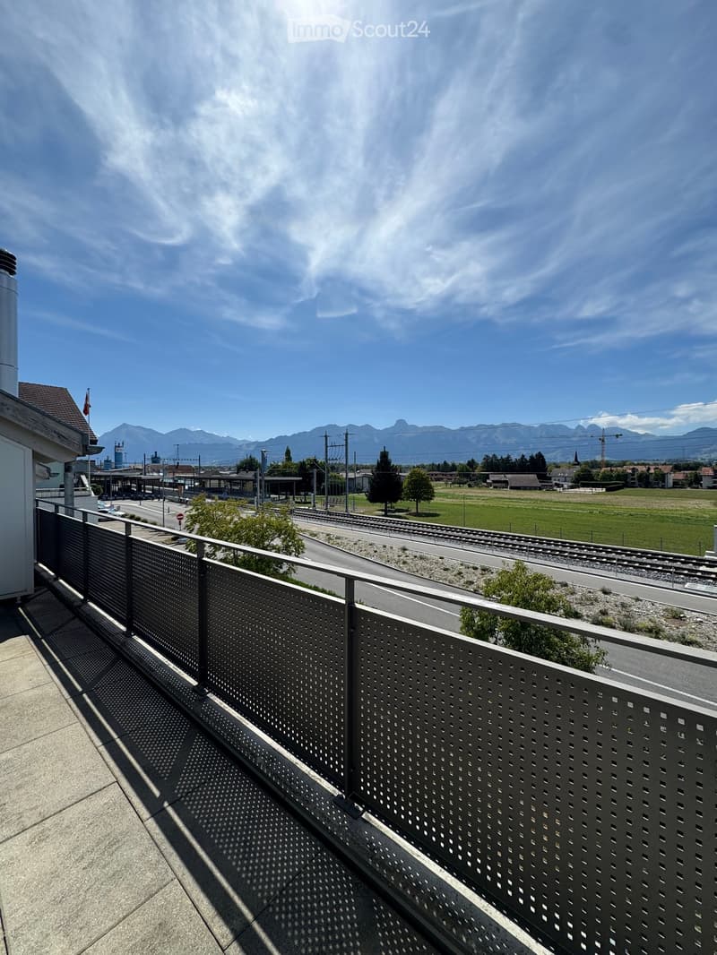 Balcony with metal railing, overlooking a train track, road, and distant mountains under a clear blue sky.