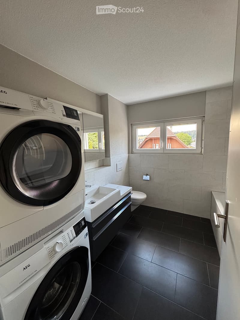 Modern bathroom with a washing machine, sink, and toilet. Features dark tiled flooring and a window with a view of a house and greenery.
